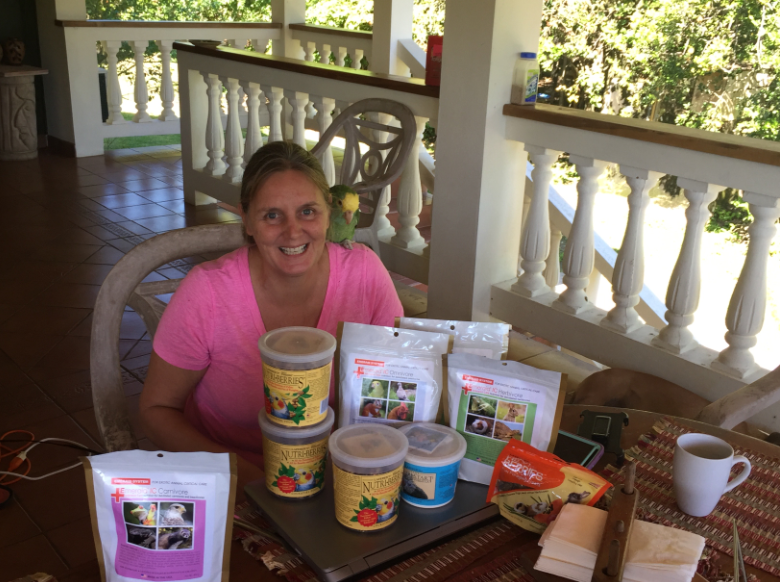 Nikki Buxton on a veranda with a parrot on her shoulder, behind a table stacked with Lafeber bird food.