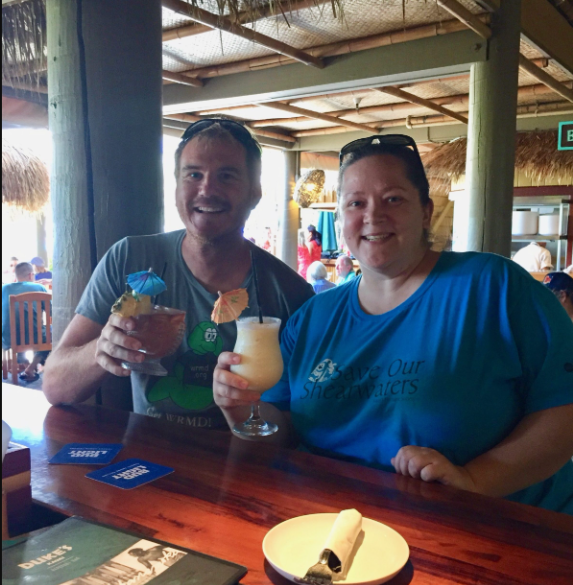 Devin and Tracy Anderson raise tropical drinks at a restaurant bar, she in a Save Our Shearwaters shirt.