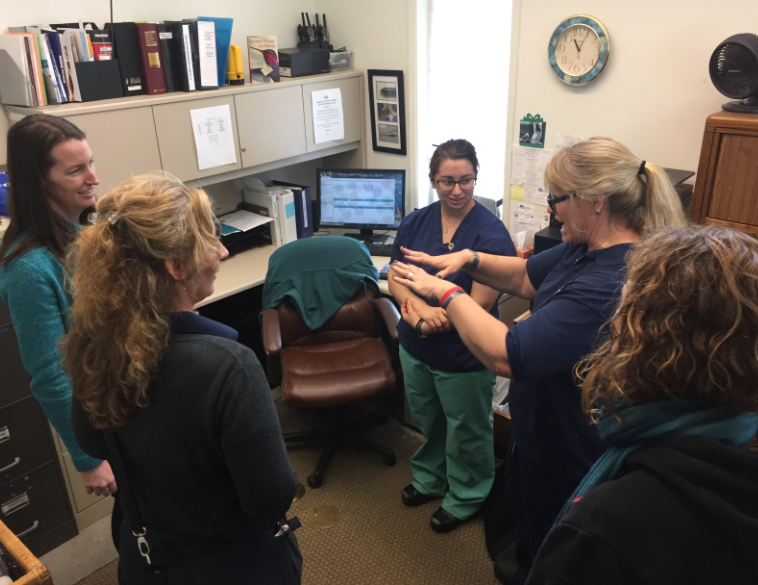 A staff member in scrubs gestures at a colleague's forearm while visitors look on in a clinic office.