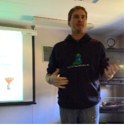 A speaker in a logo hoodie presents beside a projected slide.