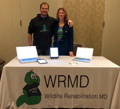 Two WRMD team members at their symposium booth, a table of laptops and tablets draped in a Wildlife Rehabilitation MD banner.