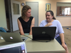 Two women talk across a table with laptops open during a working session.