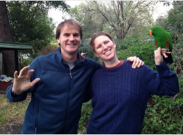 A man waving and a woman with a green parrot perched on her hand, both smiling outdoors.