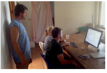 Three people gathered around a desktop computer on a long wooden table.