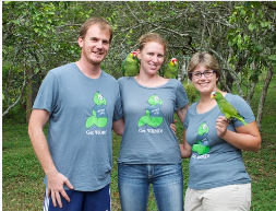 Three team members in matching WRMD T-shirts outdoors among trees, with parrots perched on their shoulders and hands.